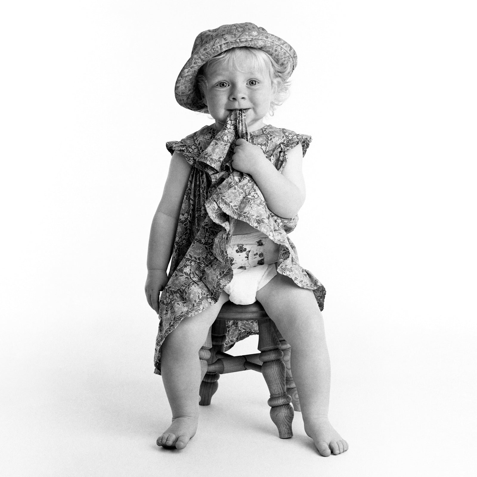 Little girl sitting on a stool, biting the hem of her dress, wearing a hat - in black and white photographed in 1996 - 