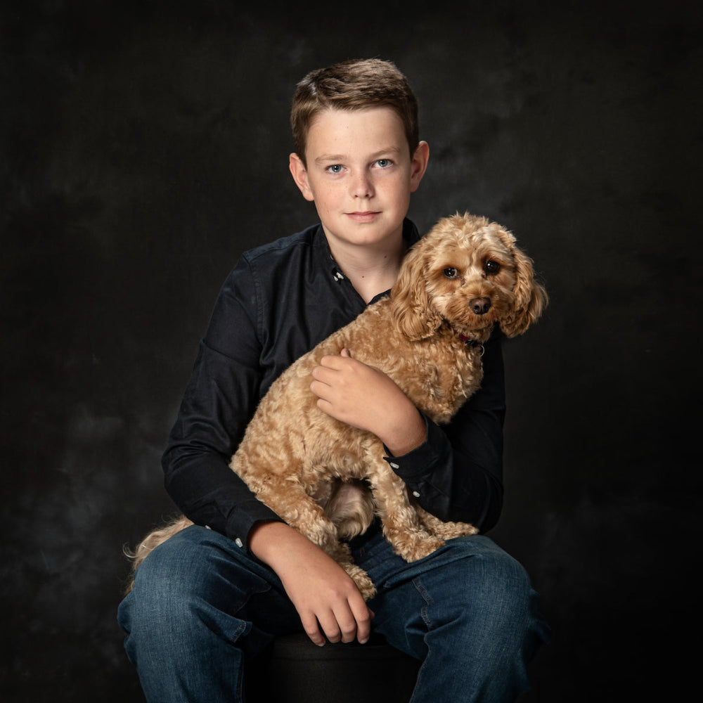 Portrait in colour of a teenage boy in dark shirt and jeans, holding his small golden Cockapoo dog against a dark background by PHOTOGENIC Dalkey