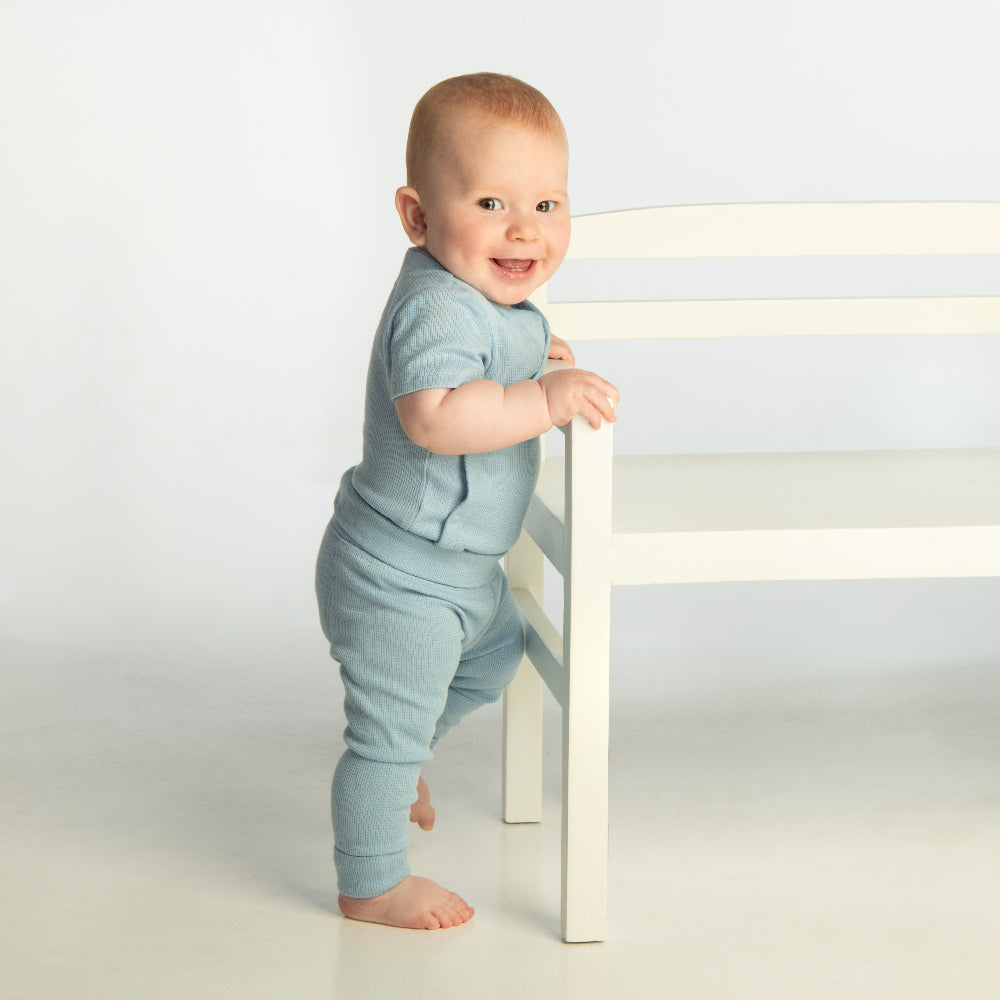 Baby in light blue outfit standing next to a chair on a white background by PHOTOGENIC Dalkey