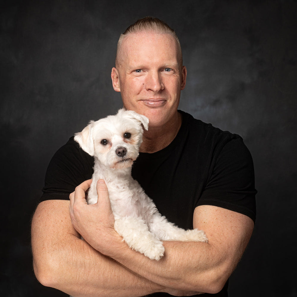 Colour Portrait of a man dressed in a black top with arms showing and holding his white Maltichon by PHOTOGENIC Dalkey