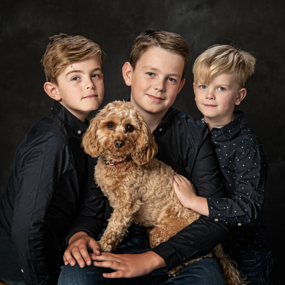Portrait in colour of three boys in dark shirts and their small golden Cockapoo dog against a dark background by PHOTOGENIC Dalkey